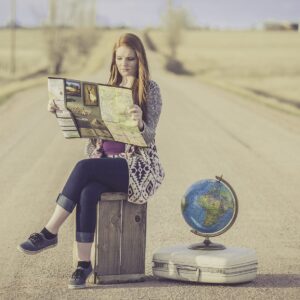 une femme assise sur sa valise au milieu de la route. Elle regarde une carte et à un globe terrestre à ses pieds